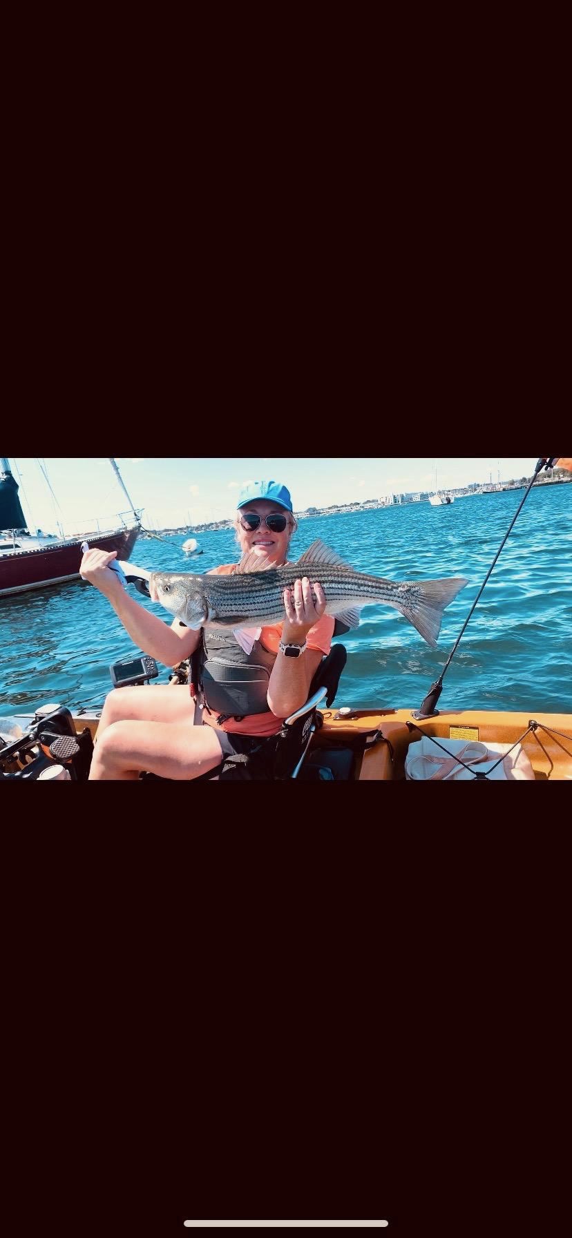 Woman kayak angler holding striped bass with sailboats in harbor background
